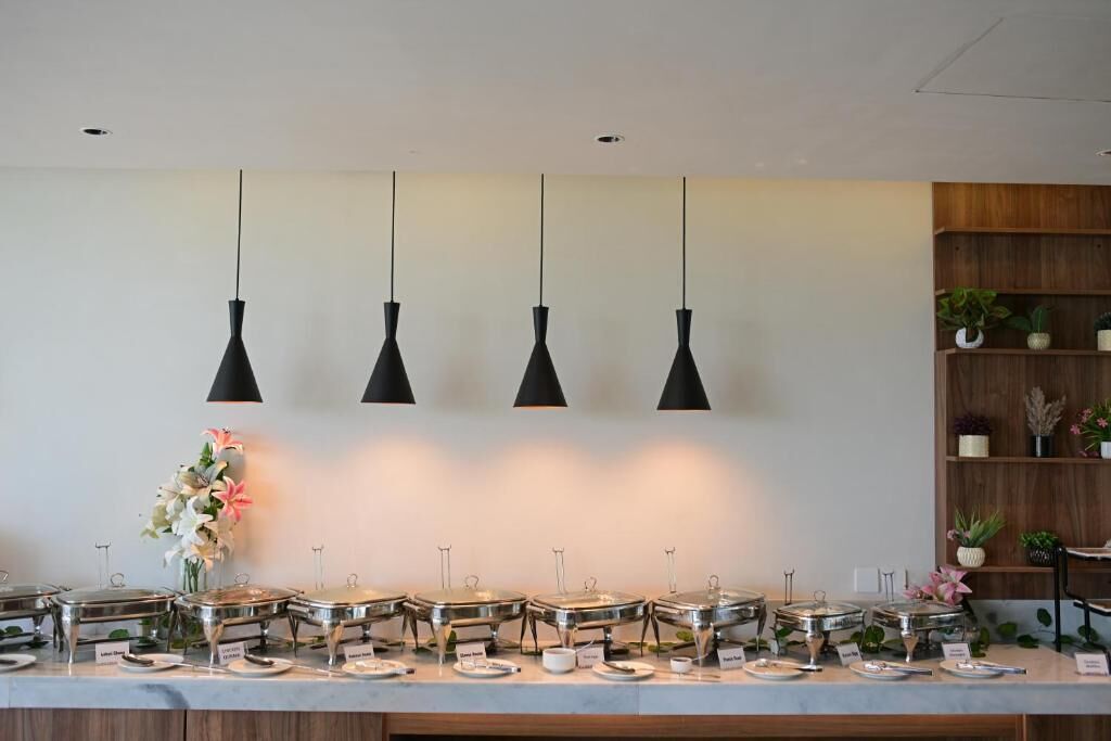A buffet setup with multiple food stations, overhead pendant lighting, and a wooden shelf with potted plants.