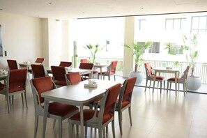 A dining area with multiple tables and chairs, large windows, and potted plants.
