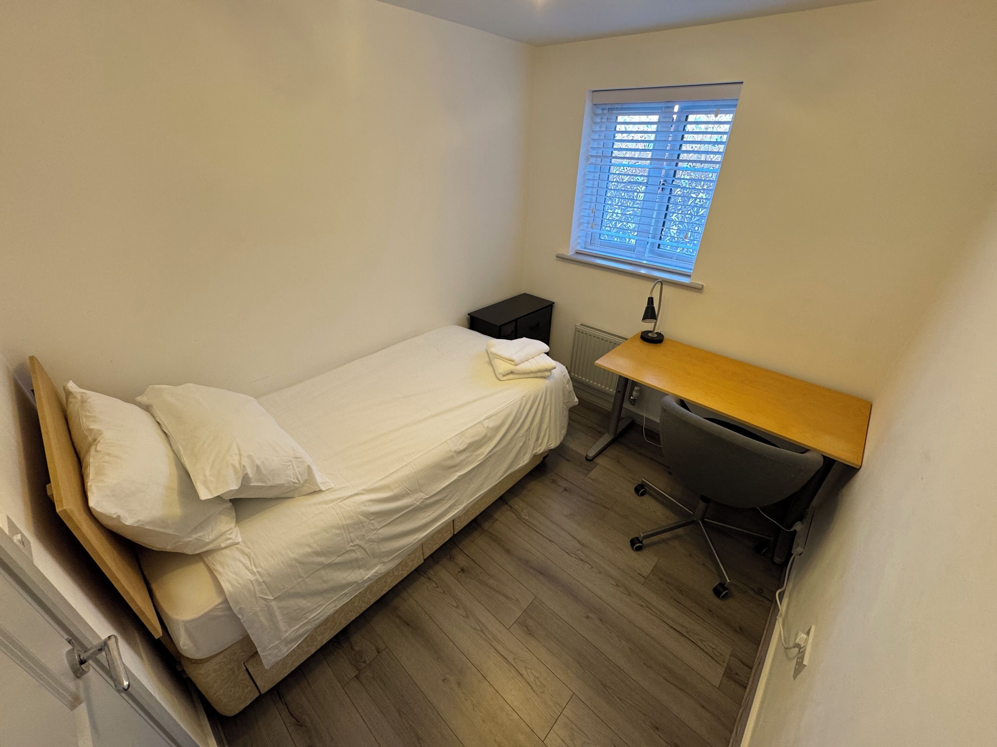 Una habitación pequeña con cama individual, suelo de madera, escritorio con silla, radiador y ventana con persianas.