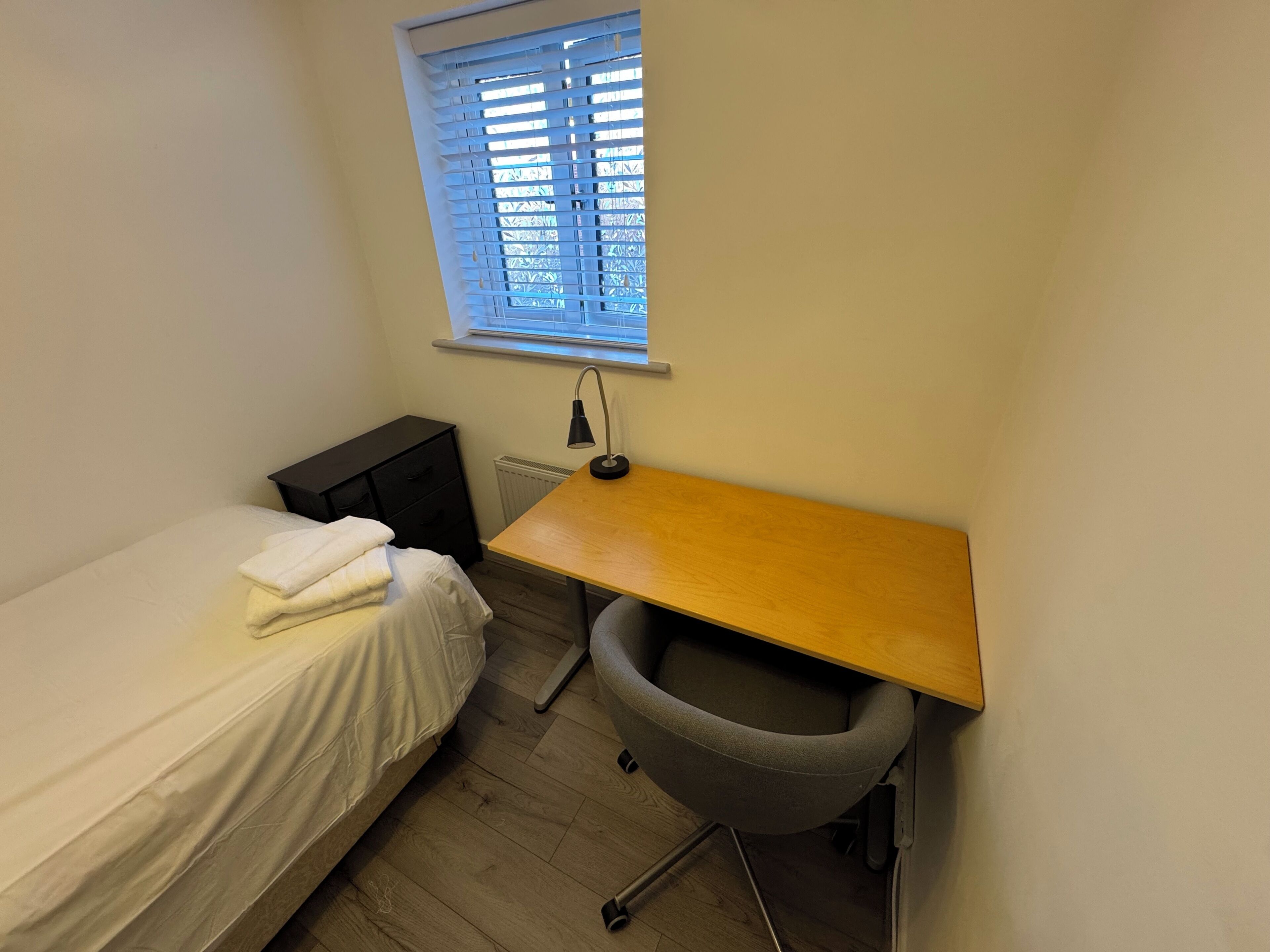 Una habitación pequeña y ordenada con una cama individual, un escritorio, una silla, un radiador y una ventana con persianas.