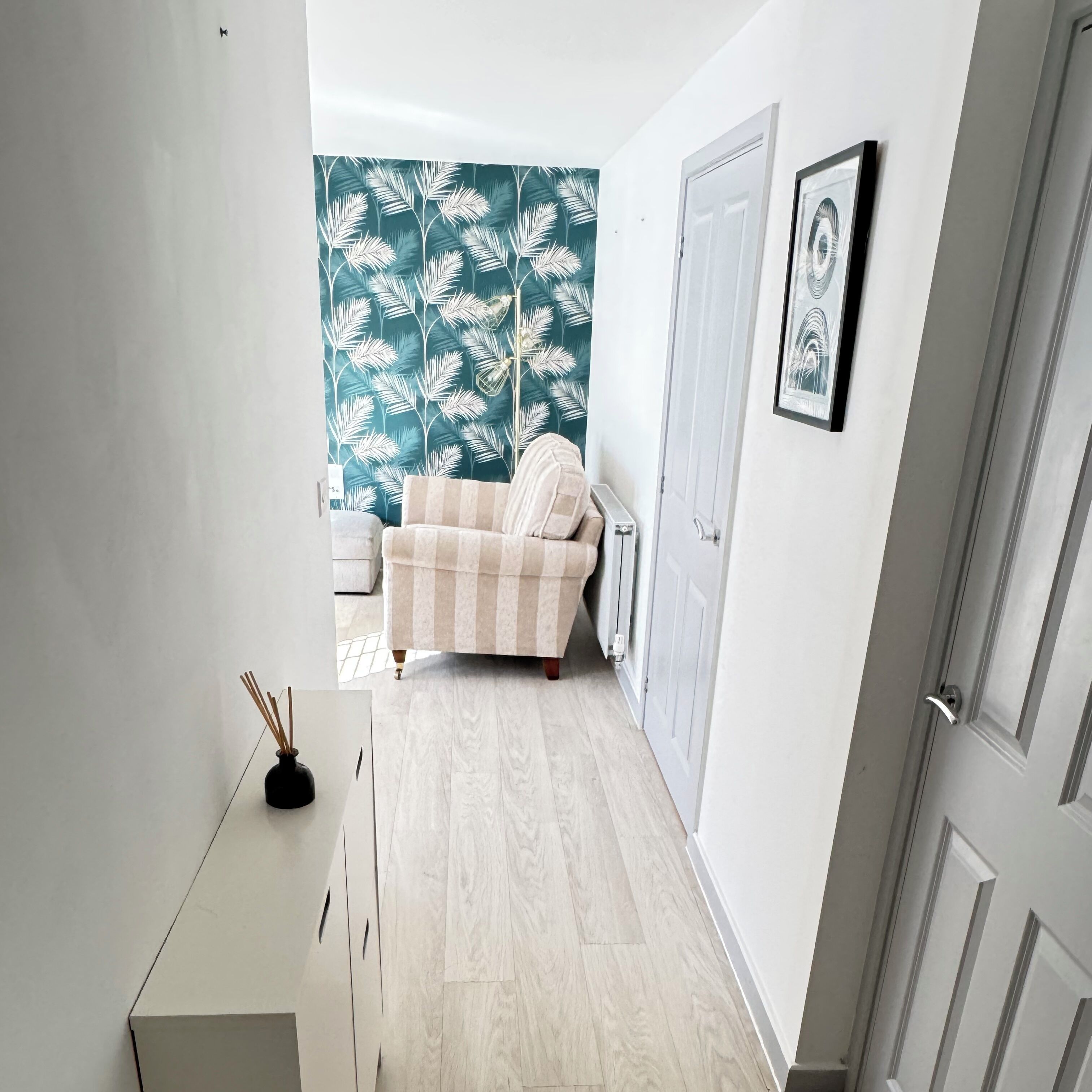 A hallway with a white cabinet, a beige sofa, a teal leaf-patterned wall, and a door with a framed picture.