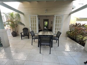 Área de varanda coberta com mesa, cadeiras, plantas em vasos e vista para o interior da casa.