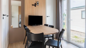 A modern room with a wooden dining table, black chairs, a flat-screen TV, and a sliding glass door leading to a balcony. - Caravan by the sea Blankenberge (Blankenberge)