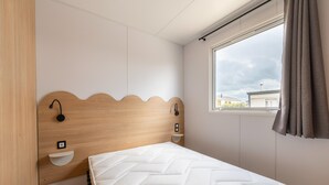 A single bed with a wooden headboard, a bedside table, a wall-mounted light, and a window with curtains. - Caravan by the sea Blankenberge (Blankenberge)