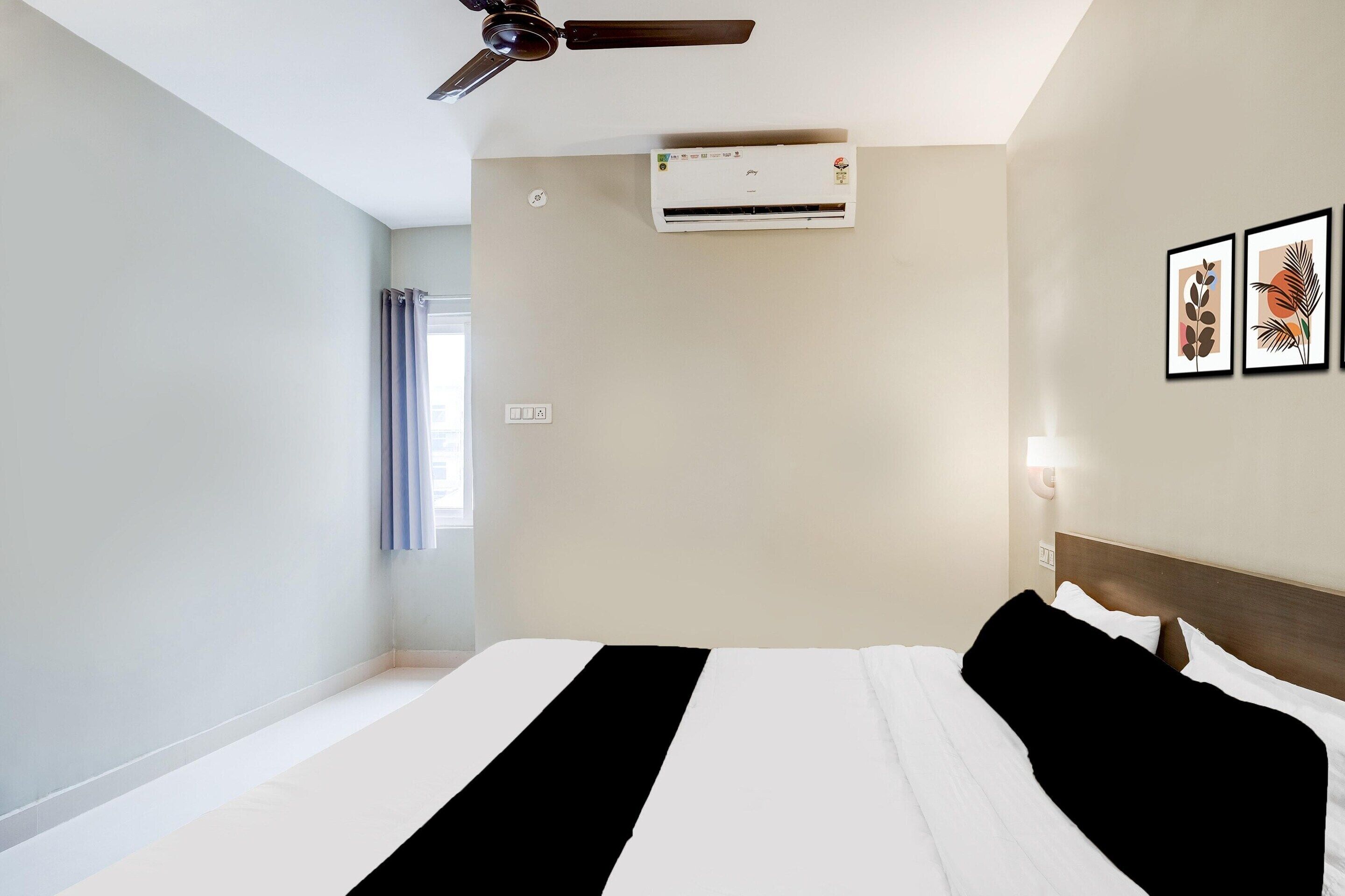A hotel room with a bed, ceiling fan, air conditioning unit, wall-mounted light, and framed artwork.