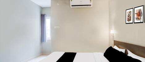 A hotel room with a bed, ceiling fan, air conditioning unit, wall-mounted light, and framed artwork.