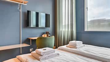 Une chambre d’hôtel avec un lit, un téléviseur fixé au mur, une fenêtre avec des rideaux et une table de chevet avec une lampe.