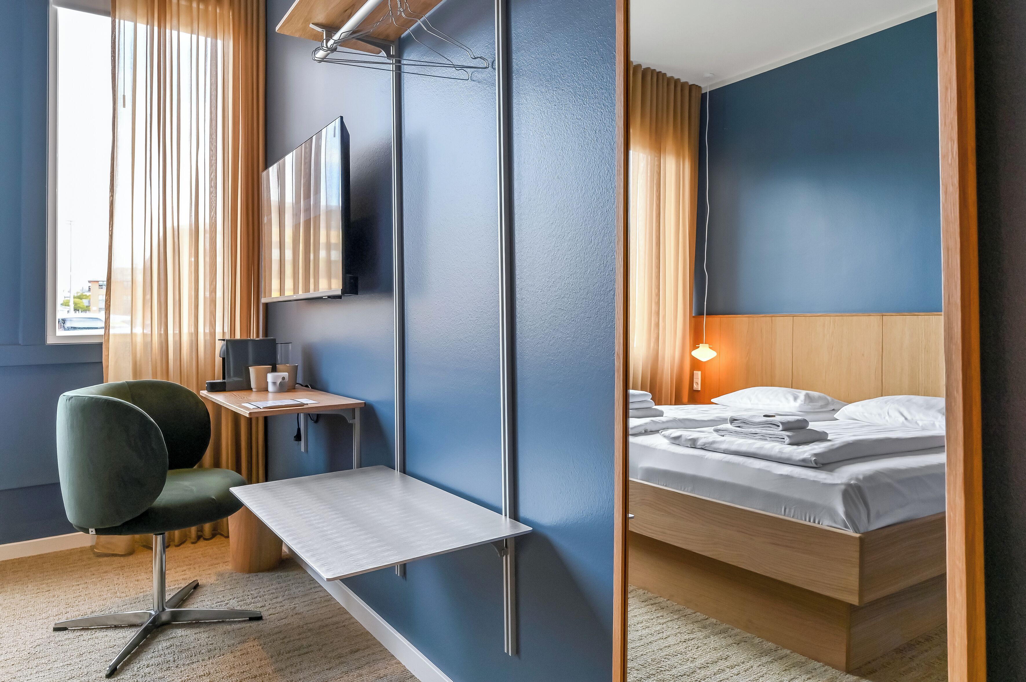 Una habitación de hotel con cama, escritorio con silla, televisor montado en la pared y ventana con cortinas.