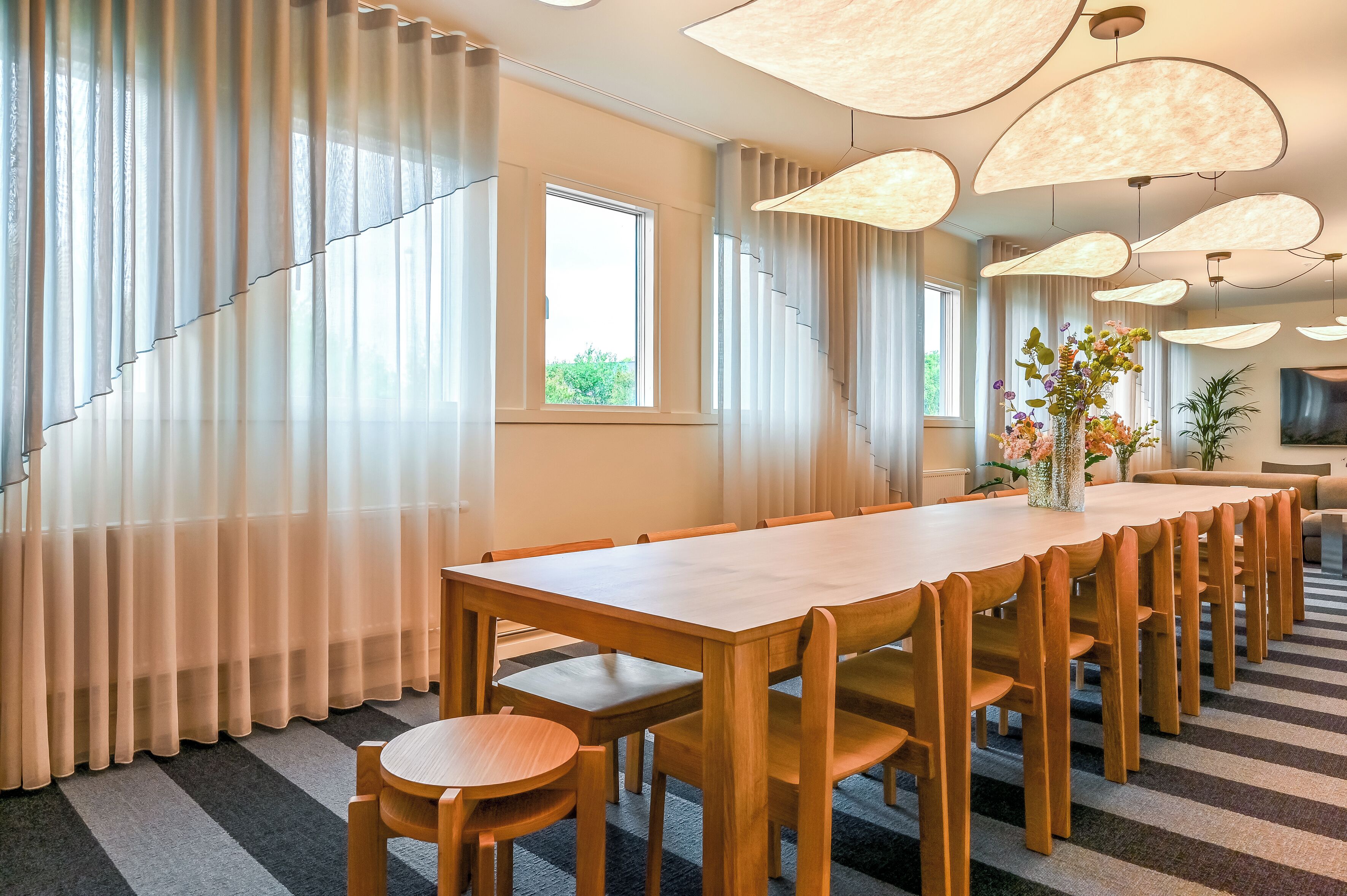 A conference room with a long wooden table, chairs, a vase with flowers, and a large window with curtains.