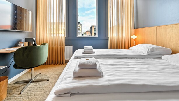 A hotel room with two beds, a desk, a chair, a window with curtains, and a lamp. - The Break Hotel (Herning)
