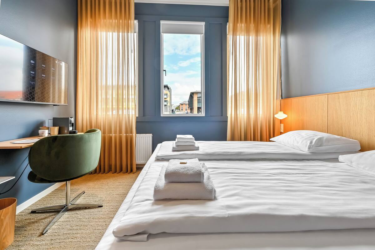 A hotel room with two beds, a desk, a chair, a window with curtains, and a lamp.
