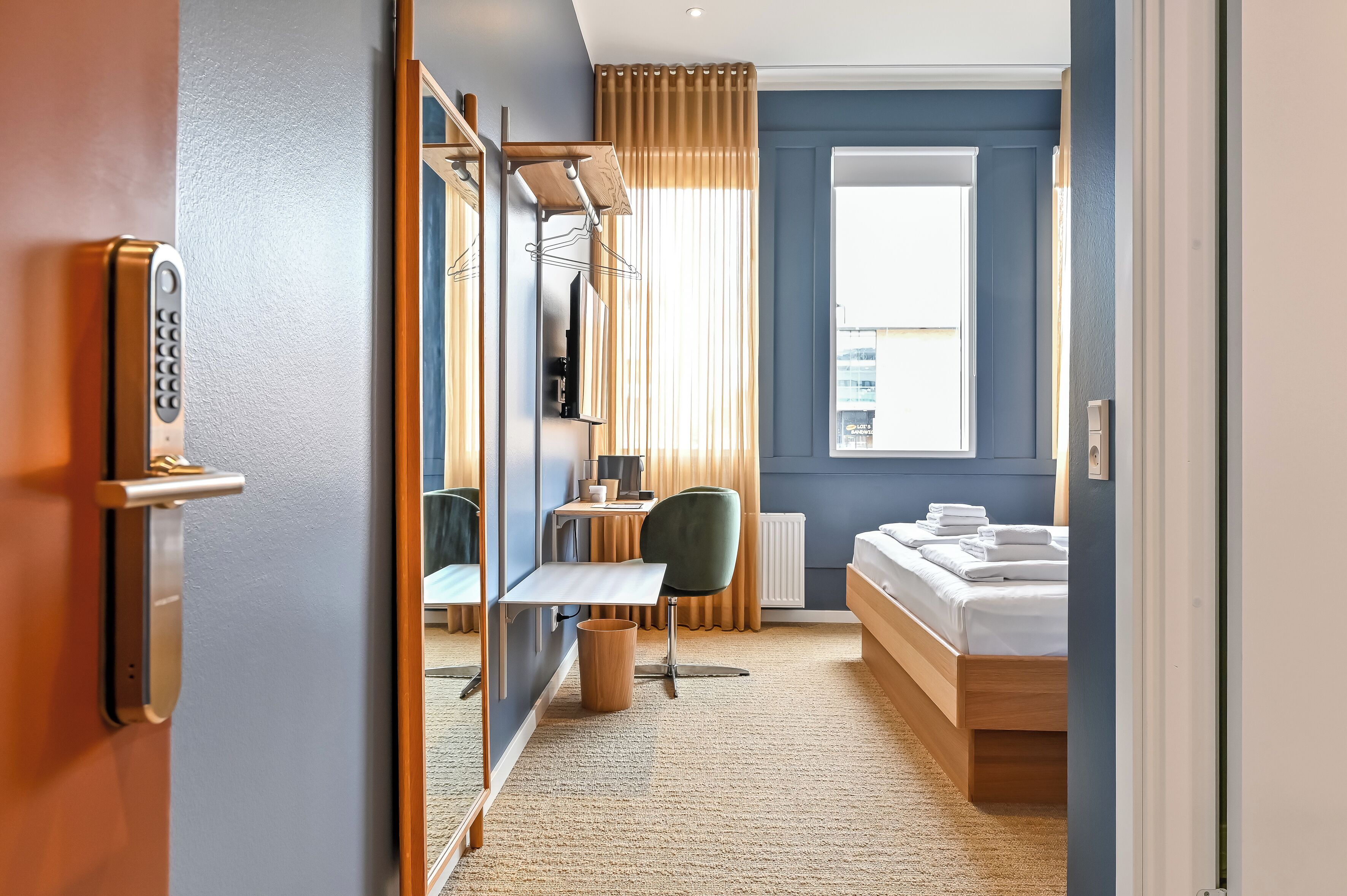 A hotel room with a bed, a desk, a chair, a mirror, a window, and a door.