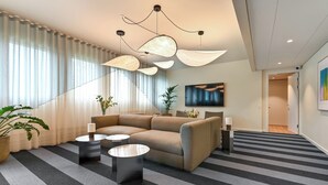 A modern living room with a sofa, two side tables, a large window with curtains, a potted plant, and a painting on the wall. - The Break Hotel (Herning)