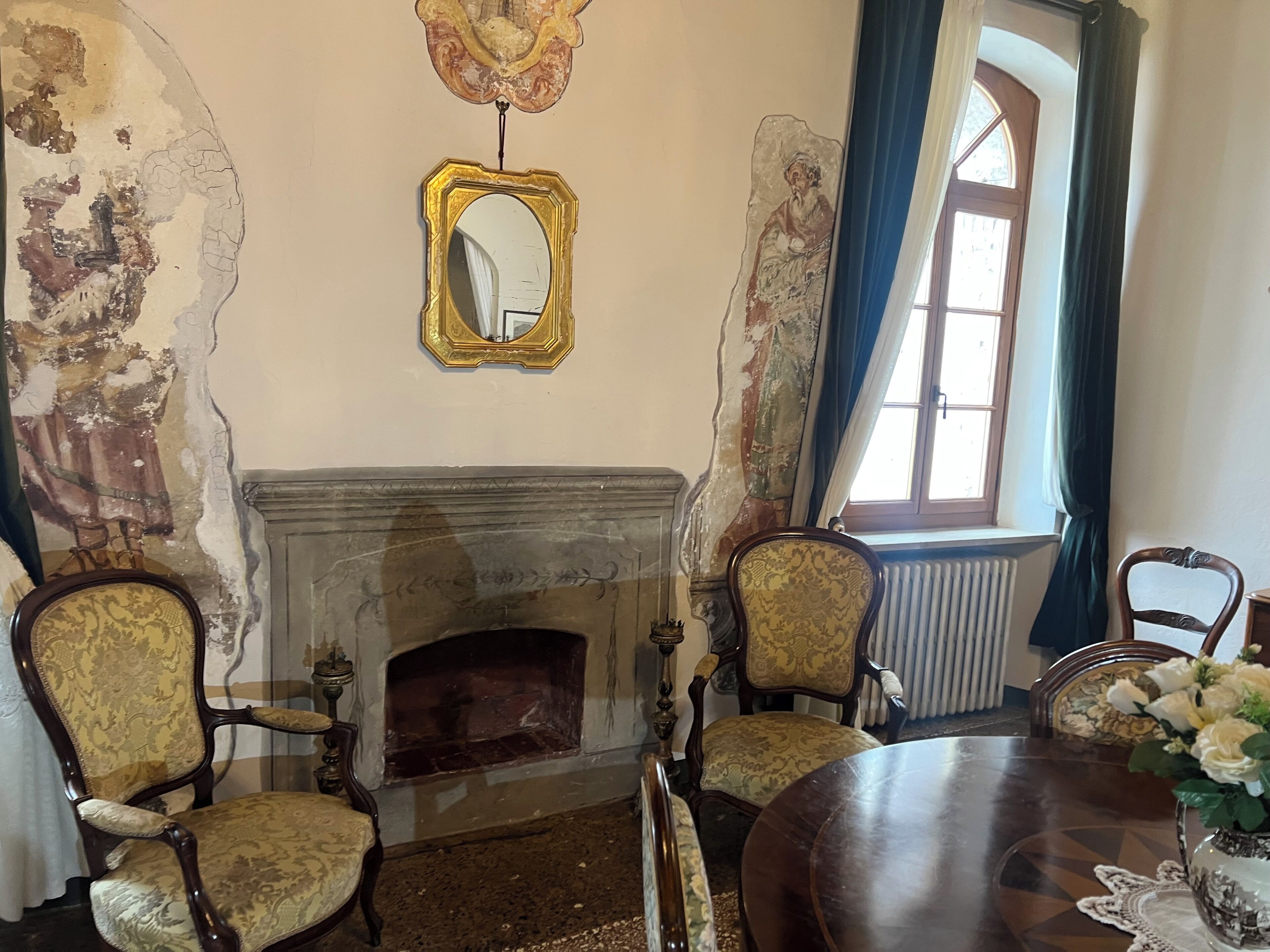 A room with a fireplace, ornate chairs, a table with a vase, and a window with curtains.