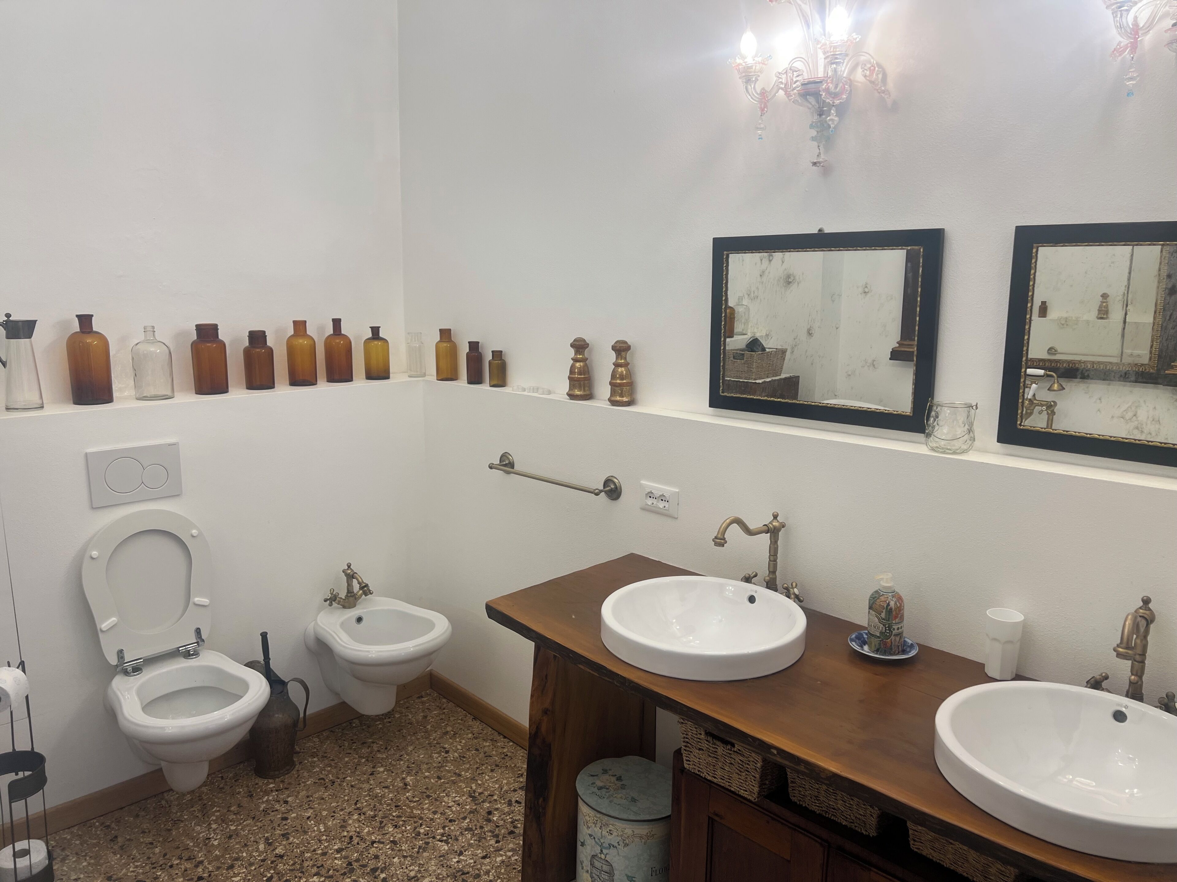 A bathroom with two sinks, a bidet, a toilet, and a row of bottles on the wall.
