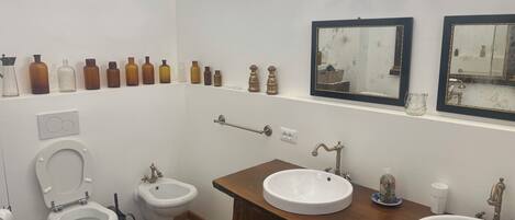 A bathroom with two sinks, a bidet, a toilet, and a row of bottles on the wall.