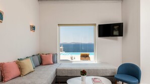 A modern living room with a sofa, a blue chair, a round table with a book and a vase, a TV on the wall, and a large window overlooking a pool and mountains. - 3-bedroom Infinity Blue villa in fantastic Sunset Views close to Mykonos Town (Mykonos)