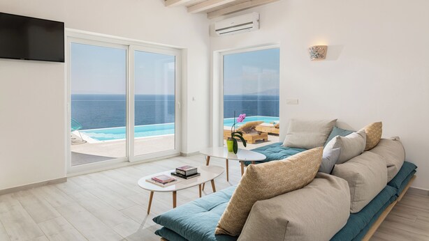 A modern living room with a large sofa, a coffee table, and a TV. There are large windows with a view of the sea. - 3-bedroom Infinity Blue villa in fantastic Sunset Views close to Mykonos Town (Mykonos)