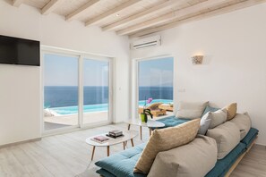 A modern living room with a large sofa, a coffee table, and a TV. There are large windows with a view of the sea. - 3-bedroom Infinity Blue villa in fantastic Sunset Views close to Mykonos Town (Mykonos)