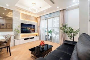 Una sala de estar moderna con un televisor de pantalla plana, un sofá de cuero negro, una mesa de centro con flores y una zona de comedor con una mesa y sillas blancas.