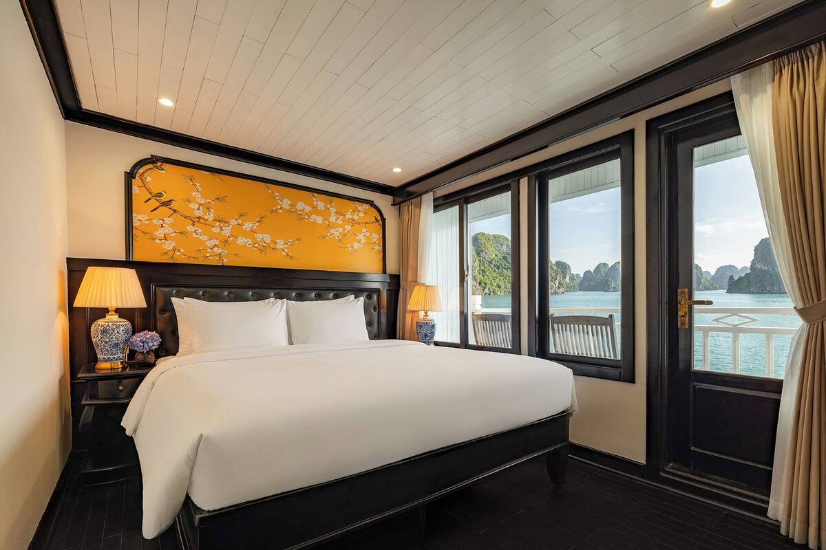 Quarto de hotel com uma cama grande, uma mesa de cabeceira com luminária, um objeto decorativo na parede e vista para o mar através de grandes janelas.
