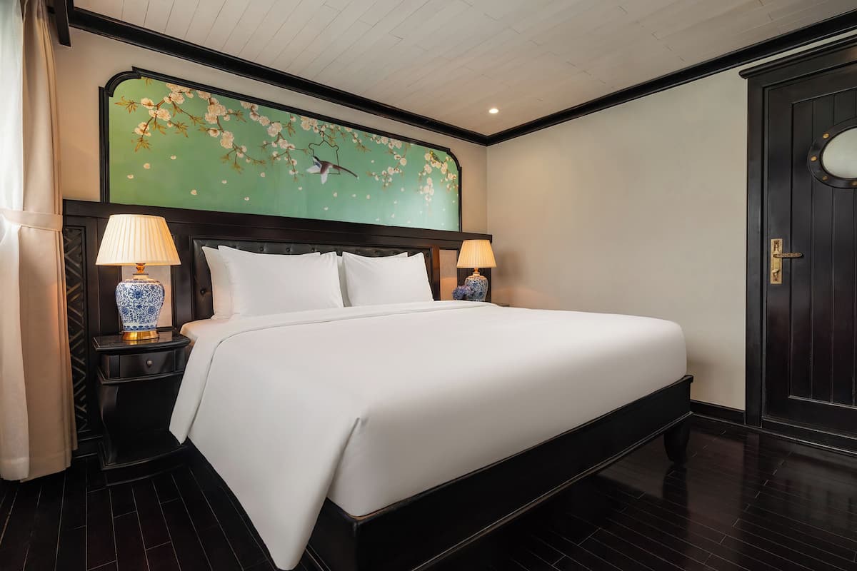 Quarto de hotel com uma cama grande, duas mesas de cabeceira com luminárias, um painel decorativo na parede e uma porta de madeira escura.