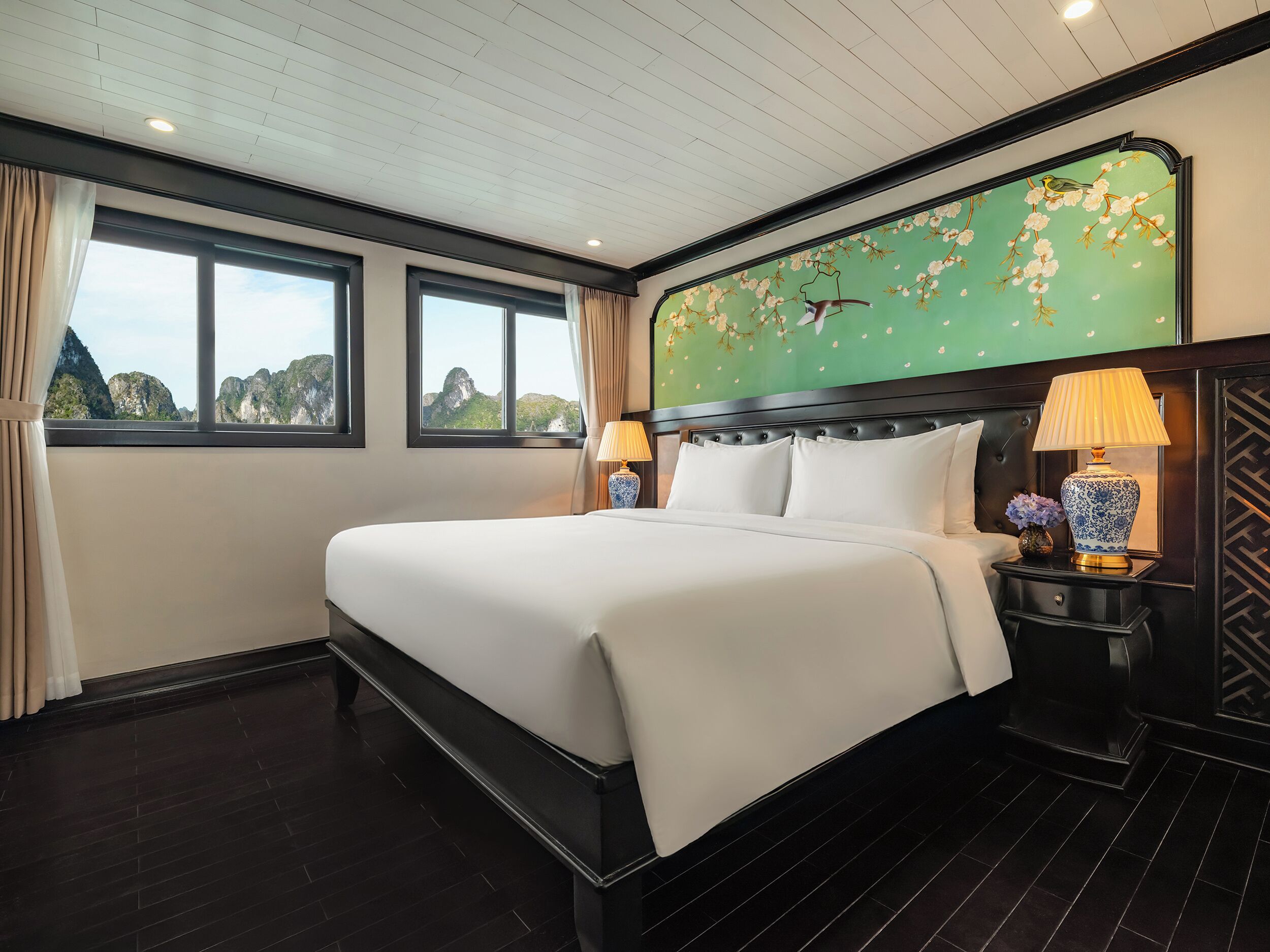 A hotel room with a large bed, two bedside lamps, a decorative wall mural, and a view of the mountains through the windows.
