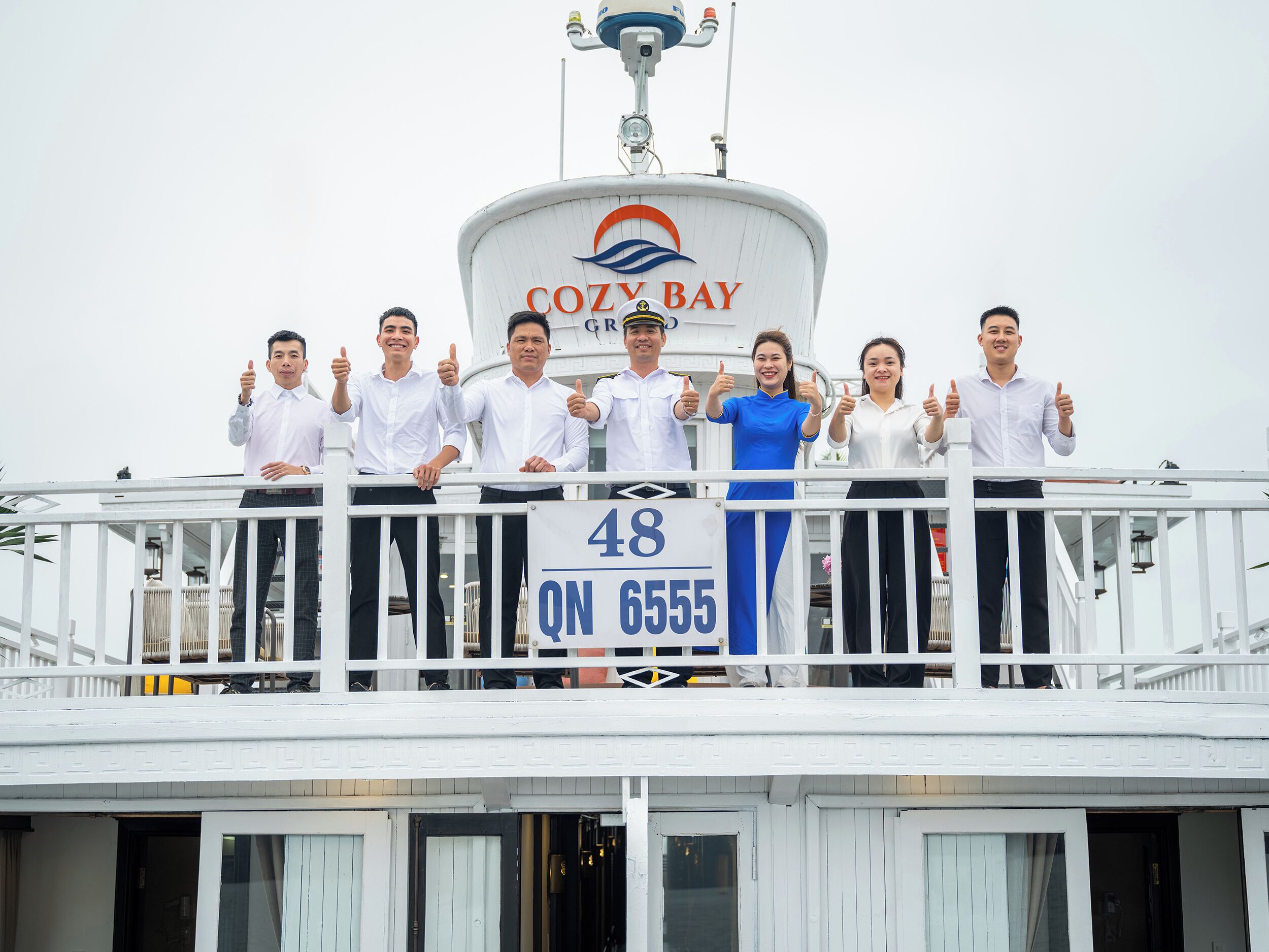 A group of people on a balcony with a sign reading 'COZY BAY' and a number '48' on a signboard.