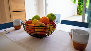 Un frutero sobre una mesa con mantel blanco, dos tazas y una ventana con follaje en el exterior.