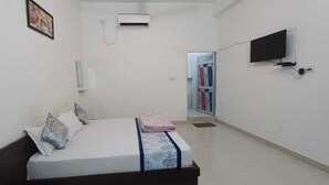 A bedroom with a bed, a TV, an air conditioning unit, and a door leading to a bathroom. - Amrit Hotel & Resort (Thanagazi)