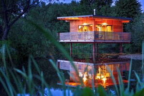 A two-story wooden cabin with a balcony, situated on stilts over water, surrounded by trees. - Besonderes Tinyhouse (Grossharthau)