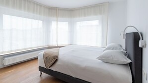 Een slaapkamer met een bed, witte gordijnen, een radiator en een wandlamp.