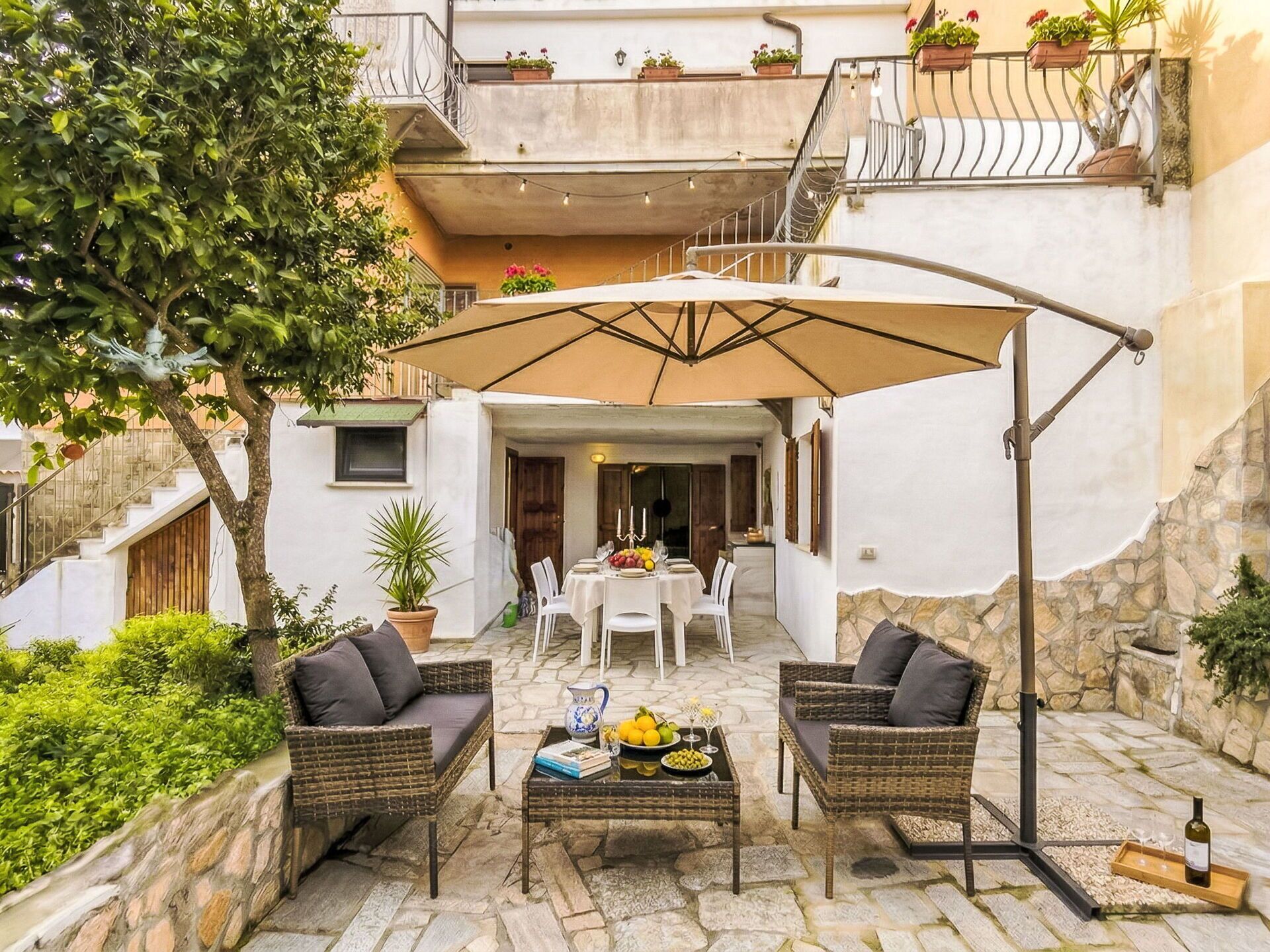 Een terras buiten met rieten meubels, een tafel met fruit en wijn, en een grote parasol.