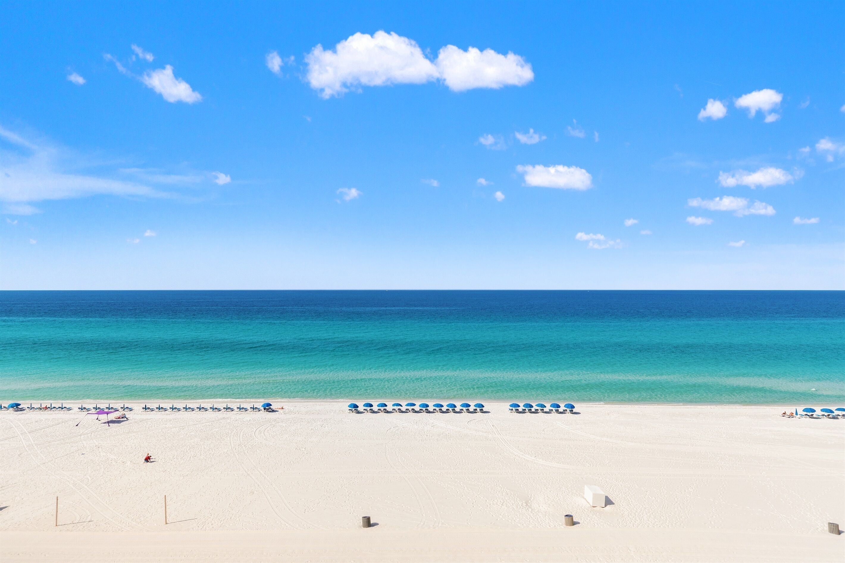 Ein Strand mit blauem Himmel, sandigem Ufer und einer Reihe blauer Sonnenschirme.