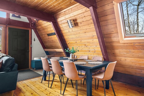 A wooden interior with a dining table set for six, a sofa, and a window with a view of trees. - Black Bear Lodge at Hunter Mountain - Hot Tub, Pellet Stove, Peace (Hunter)
