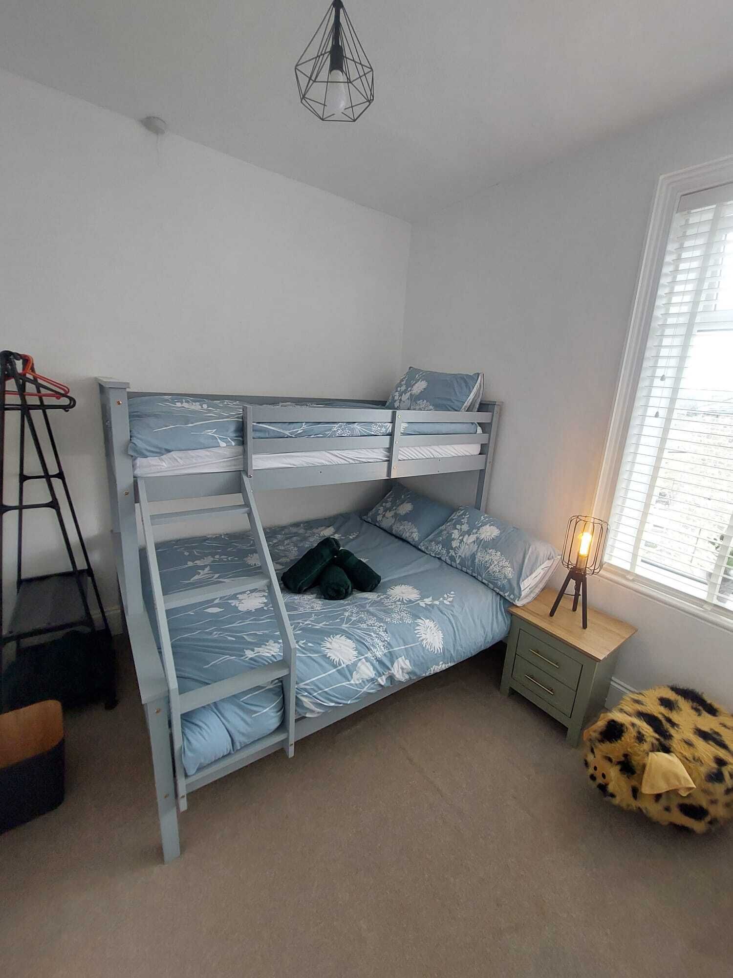 A bunk bed with a blue floral bedspread, a small bedside table with a lamp, and a leopard print ottoman.
