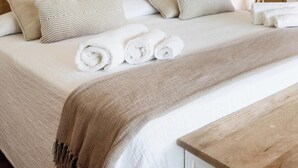 Een net opgemaakt bed met opgerolde witte handdoeken, een houten bankje en een houten dressoir in een kamer met houten vloeren.