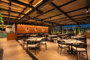 Un restaurante con techos altos, vigas a la vista y una pared de ladrillo decorativa. Las mesas están dispuestas para comer, y se aprecia una vista de vegetación al exterior.