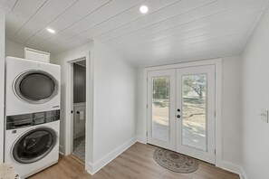 Une buanderie équipée de deux machines à laver et de deux sèche-linge à chargement frontal superposés, une porte donnant sur un espace extérieur abrité et un petit tapis sur le sol.