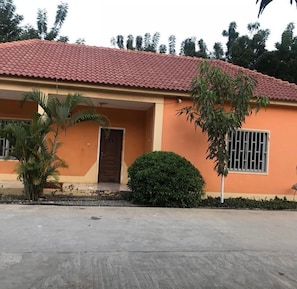 A single-story building with a red-tiled roof, a front door, and a small garden area with a palm tree and a bush.