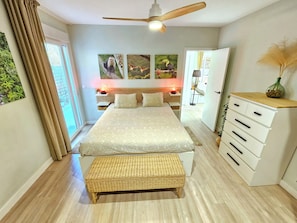 A bedroom with a bed, a bedside table, a lamp, a ceiling fan, a window with curtains, and wall art.