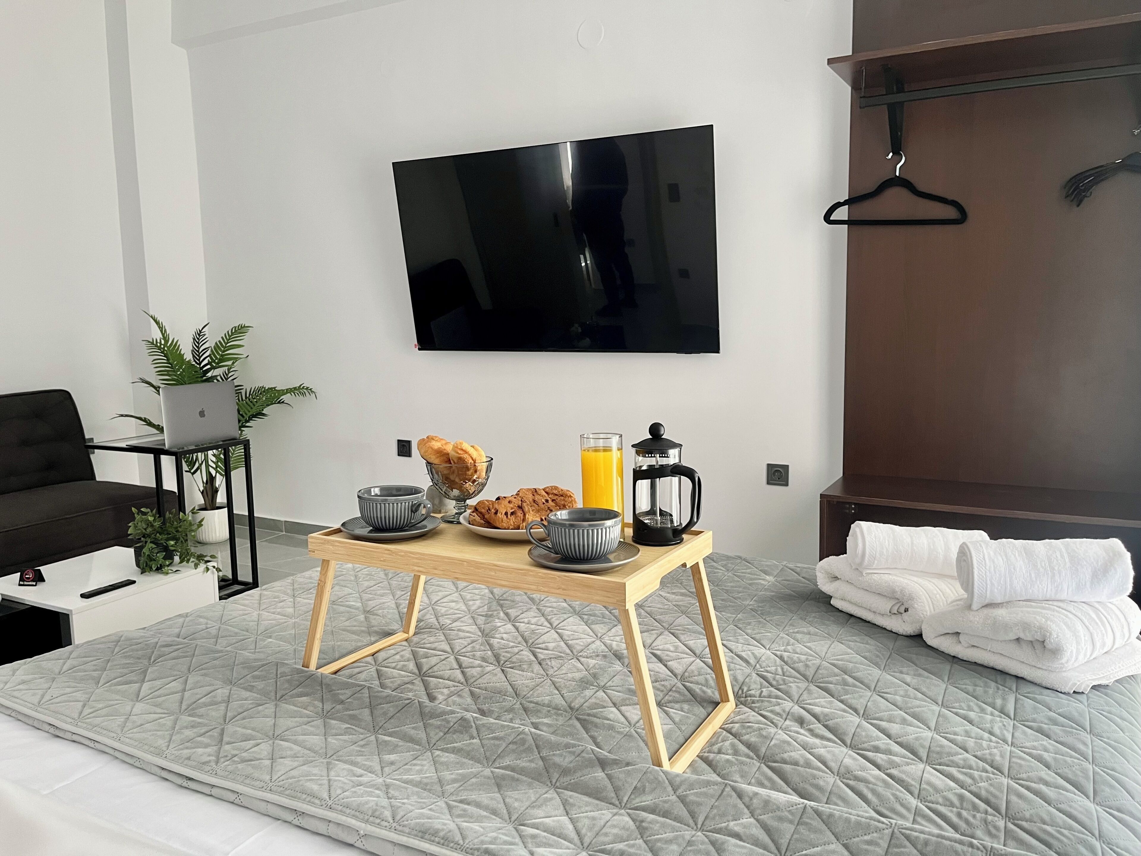 Un dormitorio ordenado con cama, una mesita de noche con artículos de desayuno, un televisor montado en la pared y un armario.