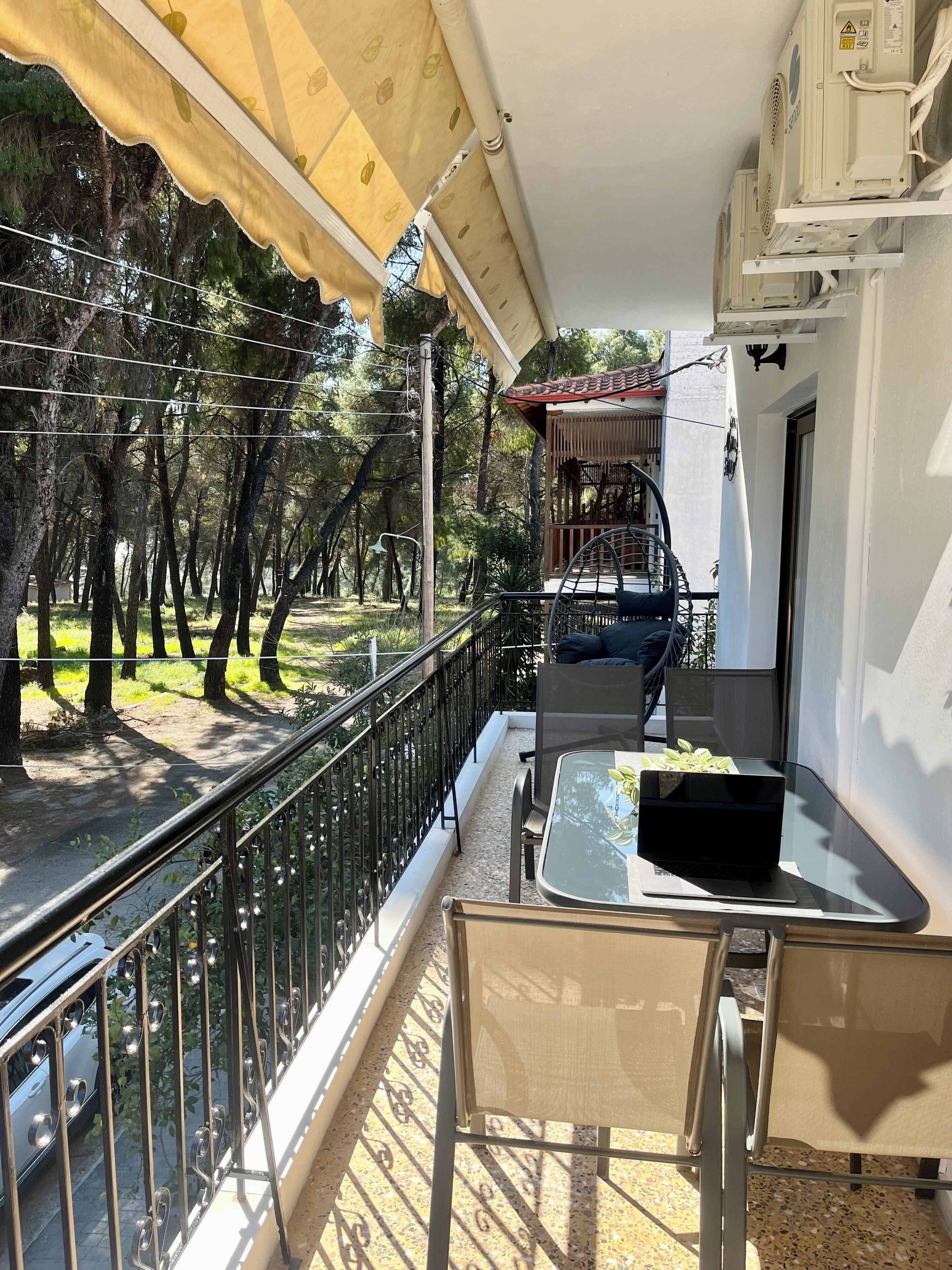 Ein Balkon mit Tisch, Stühlen und einem Laptop. Ein Geländer, Blick auf Bäume und ein Gebäude im Hintergrund sind zu sehen.