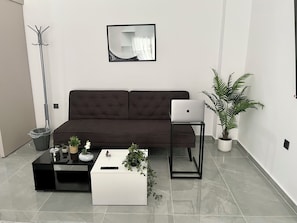 Una sala de estar moderna con un sofá marrón oscuro, una mesa de centro blanca, una computadora portátil sobre un soporte, una planta en maceta y una lámpara de pie.