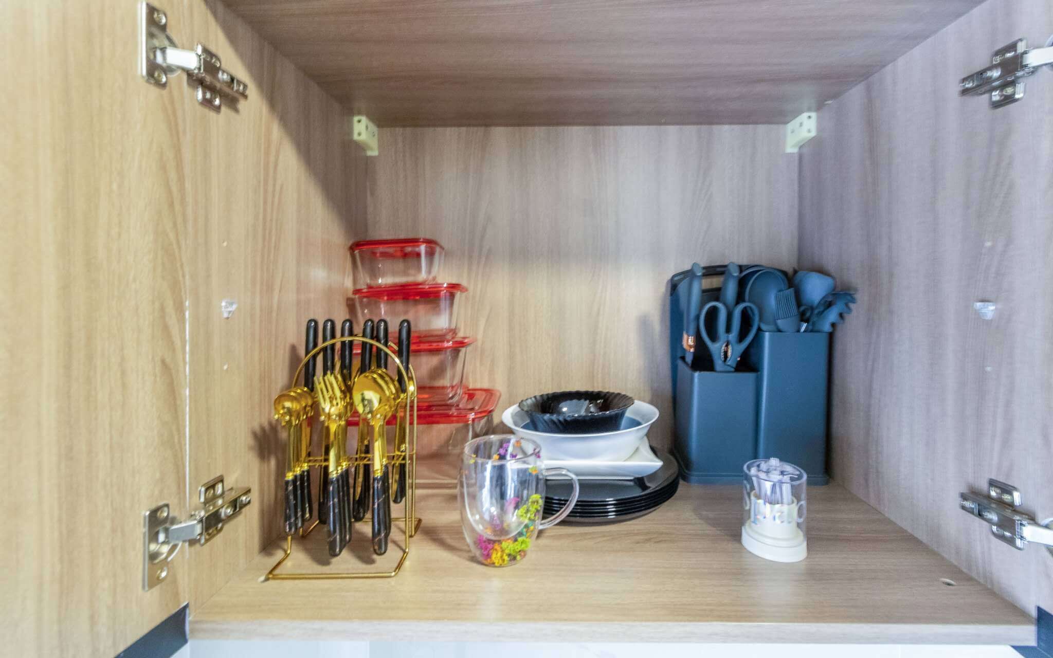 Ett träskåp med metallgångjärn, innehållande ett set verktyg med gyllene handtag, röda och genomskinliga plastbehållare, en vit mugg med färgglada föremål, en svartvit tallrik och en blå hållare för köksredskap.