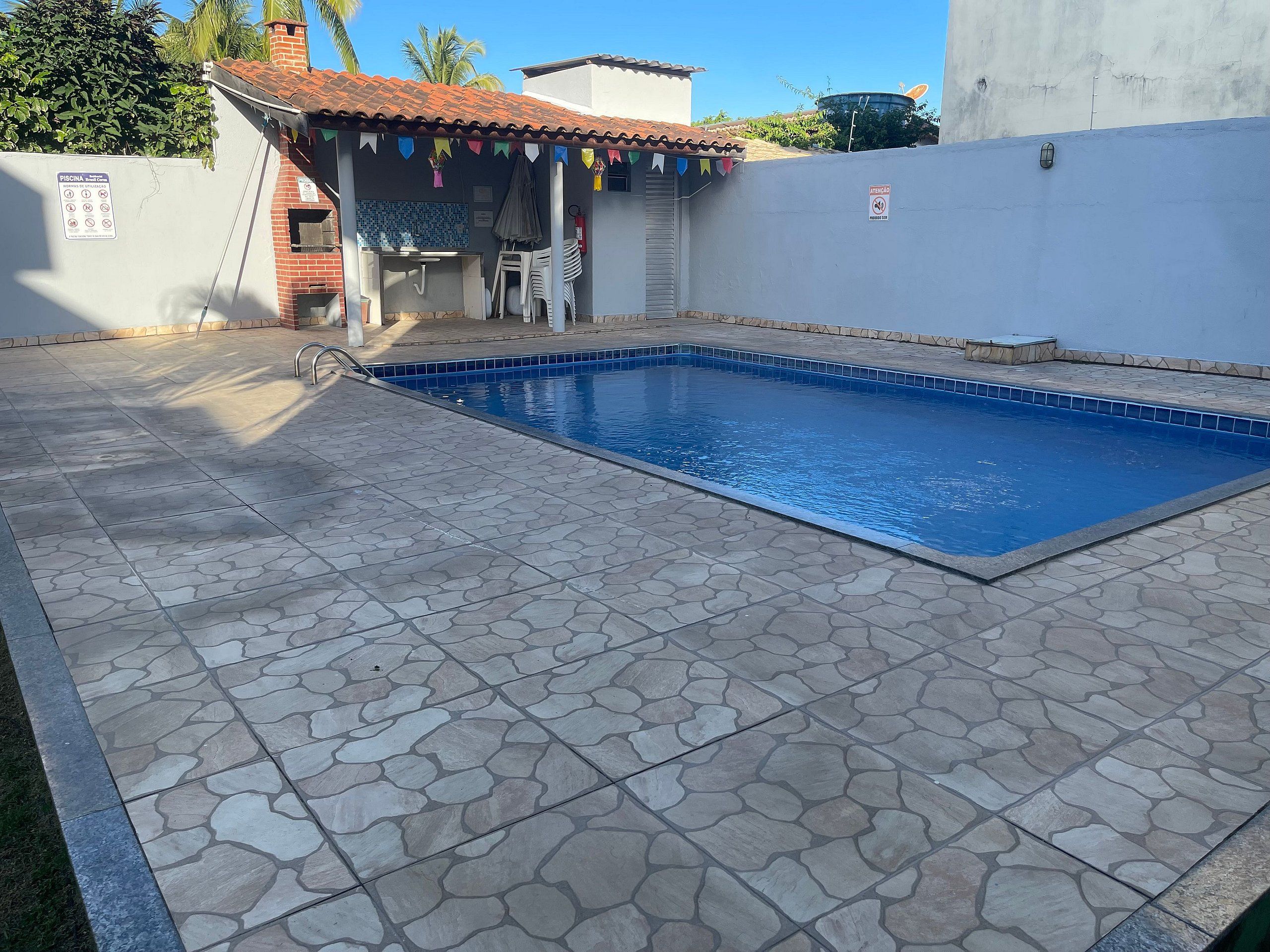A swimming pool with stone tiles, a small covered area with chairs, and a wall with a sign.