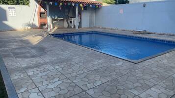 A swimming pool with stone tiles, a small covered area with chairs, and a wall with a sign.