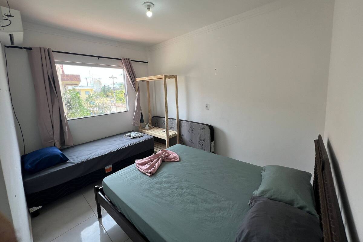 Una habitación pequeña y sencilla con cama, banco, ventana y lámpara en el techo.