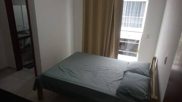 A single bed with a green bedspread in a room with a window, a door, and a mirror.