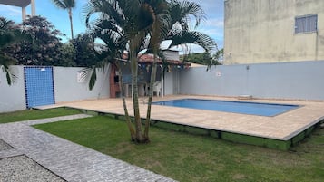 Uma piscina com deck revestido, área gramada e um edifício com porta azul.